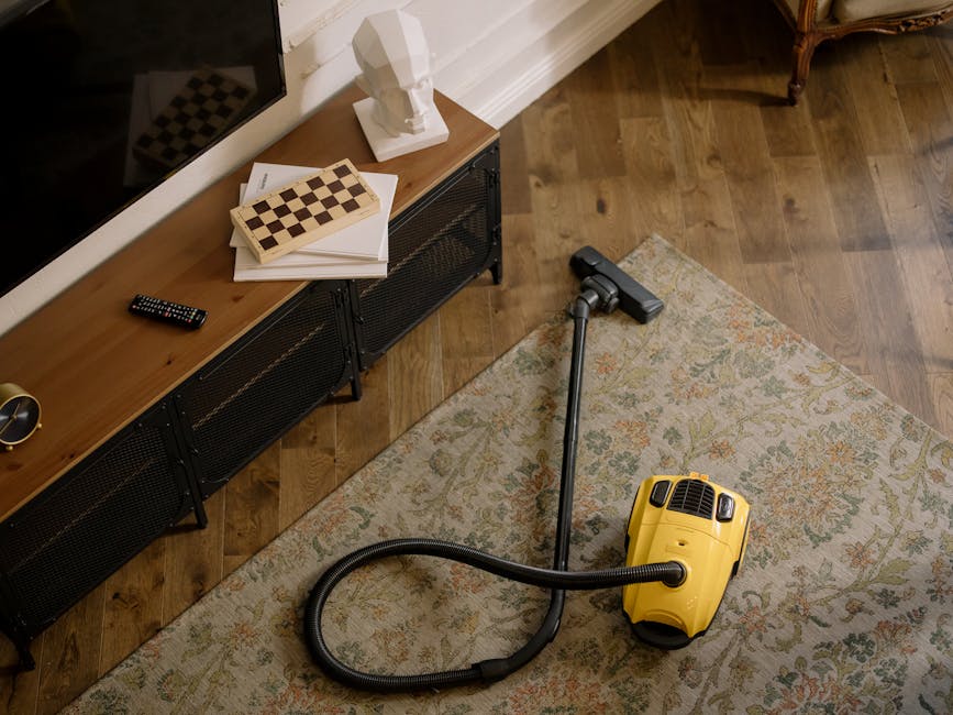 A person in a casual beige jacket and blue jeans using a yellow vacuum cleaner with a long hose to perform deep cleaning or surface cleaning on a patterned carpet in a living room. The room features w